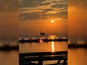 A stunning sunset over the ocean, with reflections shimmering on the water and an oil rig silhouetted against the vibrant sky — a perfect backdrop for evening boat trips.
