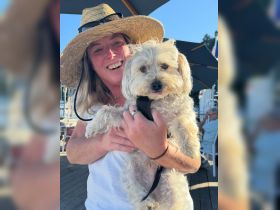 A woman smiles while holding a small dog, enjoying a sunny day at a marina. The background is not clearly defined.