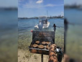 A close-up of burgers sizzling on a grill, with a boat in the background, showcasing a perfect barbecue setup for a fun day on the water.
