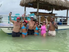 A lively group celebrates in the water next to a pontoon boat, enjoying drinks and fun under the sun — the perfect backdrop for a memorable day on the water.