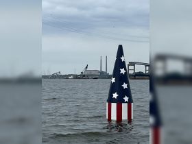 An unclear image featuring a buoy with a patriotic design in the foreground and industrial structures in the background, set against a cloudy sky.