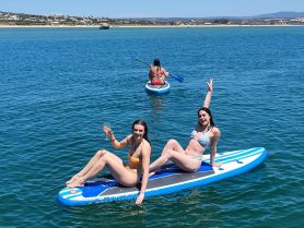 Two friends enjoy paddleboarding in clear waters, celebrating summer with drinks in hand — a perfect day for fun on the water.