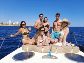 A lively group enjoys a sunny day on a yacht, celebrating with cocktails and smiles against a beautiful coastal backdrop.