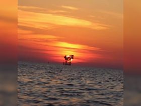 A sunset view over the water with an industrial structure silhouetted against the vibrant sky, creating a serene yet ambiguous scene.