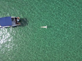 An aerial view captures a person swimming in crystal-clear waters beside a boat, showcasing the perfect summer day on the water.