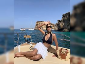 A stylish woman enjoys the sun on a boat, with stunning cliffs and turquoise waters in the background — perfect for a luxurious day on the water.