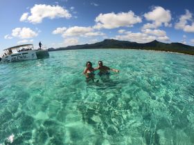 A couple enjoys a refreshing swim in crystal-clear waters with a catamaran in the background, perfect for island hopping adventures.