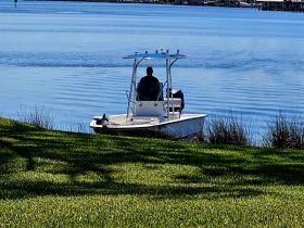 A serene view of a center console boat docked by the shore, with a person seated at the helm, surrounded by calm waters and lush greenery.