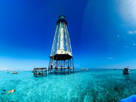 A vibrant scene featuring snorkelers exploring the crystal-clear waters near a lighthouse, with boats dotting the picturesque landscape — a perfect spot for aquatic adventures.