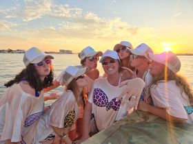 A lively group of friends celebrates at sunset, showcasing fun hats and playful poses — perfect for bachelorette parties or memorable outings on the water.
