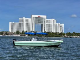 A sleek speedboat named 'Wild Tuna' is anchored in front of the RIU Hotel, showcasing a vibrant turquoise color against the backdrop of a sunny sky.