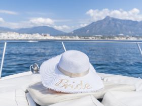 A stylish bride's hat rests on the deck of a boat, set against a stunning coastal backdrop — perfect for bridal celebrations on the water.