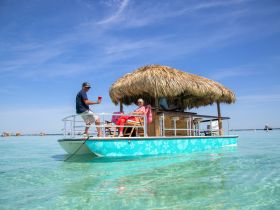 A lively group enjoys drinks aboard a tiki boat, soaking up the sun in crystal-clear waters — perfect for a fun day on the water.