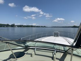 A clear view from the front of a boat cruising on calm waters, showcasing a serene landscape with trees lining the shore under a bright blue sky.