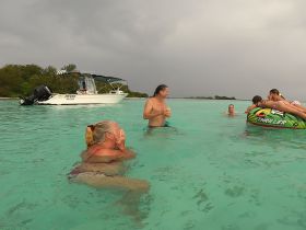 A lively group enjoys a fun day in the turquoise waters, with some swimming and others on a tube, while a boat is anchored nearby — perfect for family adventures and water activities.