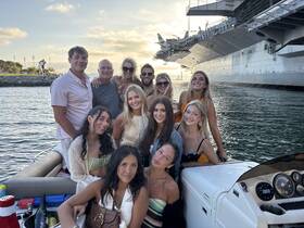 A lively group of friends poses for a photo on a boat, enjoying a beautiful day on the water with a stunning backdrop of a ship.