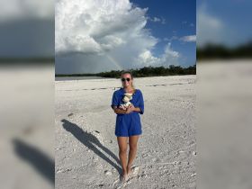 A woman stands on a sandy beach holding a collection of shells, with a rainbow visible in the background — a picturesque moment capturing the beauty of nature.