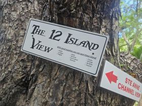 A sign indicating 'The 12 Island View' with directions to various islands, set against a tree backdrop — a scenic spot for island spotting.