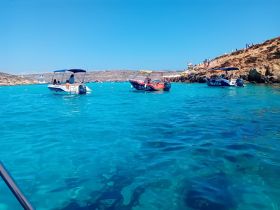 A vibrant scene featuring boats in the stunning turquoise waters of Malta, with people enjoying the beach and coastline — a perfect snapshot of summer adventures on the water.