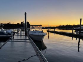 A serene sunset view of a boat docked at the marina, showcasing the tranquil waters and the silhouette of surrounding trees.