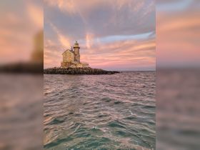 A stunning sunset view of a lighthouse standing on rocky shores, surrounded by tranquil waters — a picturesque scene perfect for coastal adventures.