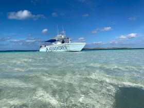 A boat named 'Aquashores' is anchored in crystal clear waters, with guests enjoying the beautiful beach scenery — a perfect setting for island adventures.