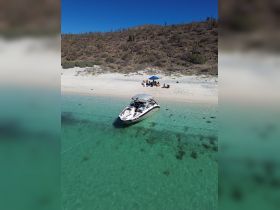 A lively group enjoys a beach day under an umbrella, with a boat anchored nearby in crystal-clear waters — perfect for a sandbar stop or a fun day out on the water.
