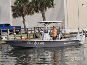 A man stands on the deck of a Blazer Bay boat named 'Fear Knot,' preparing for a fishing trip in calm waters — perfect for anglers seeking adventure.