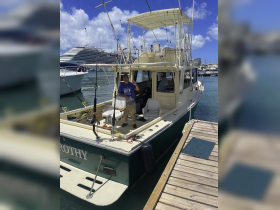 A man poses on the deck of a fishing boat named 'DROTHY' at the dock, showcasing the vessel's features against a backdrop of other boats and a cruise ship.