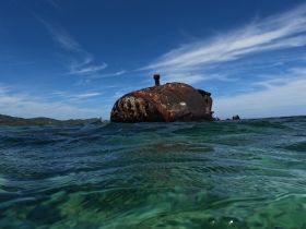 An unclear image of a submerged object in shallow water, possibly a wreck or debris, with a scenic backdrop of hills and a clear sky.