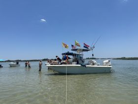 A lively group enjoys a sandbar stop, with people swimming and socializing around the boat — a perfect day for fun in the sun.