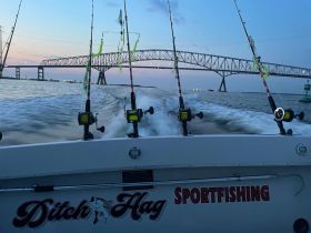 A sportfishing boat named 'Ditch Hag' speeds through the water at sunset, with fishing rods ready for action and a bridge in the background, showcasing the thrill of fishing adventures.