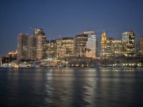 A blurry night view of a city skyline with illuminated buildings reflecting on the water, creating an abstract urban landscape.
