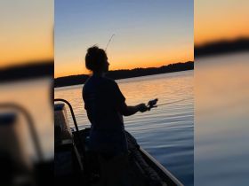 A silhouette of a person fishing at sunset, capturing the serene beauty of a tranquil evening on the water — perfect for fishing enthusiasts and nature lovers.