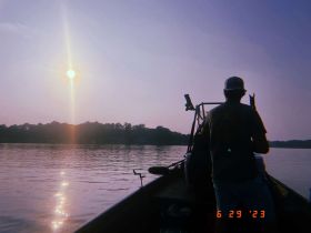 A serene sunset view from a boat with a person fishing, capturing the tranquil beauty of the water and nature.