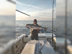A proud angler holds up a large striped bass aboard a fishing boat during a beautiful sunset — a perfect moment for fishing enthusiasts.