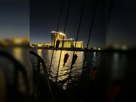 A nighttime view from a boat showing fishing rods and a hotel in the background, with reflections on the water creating a serene atmosphere.