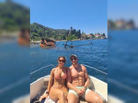 A couple enjoys a sunny day on their boat, with a scenic backdrop of an old shipwreck and lush greenery — perfect for adventure seekers and beach lovers.