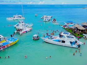 A vibrant scene at a sandbar with multiple boats and groups of people enjoying swimming and festivities in the clear turquoise waters — a perfect day for a boat party.