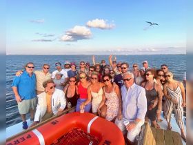 A lively group of friends gathers on a boat, enjoying a fun celebration with drinks and smiles against a beautiful ocean backdrop.