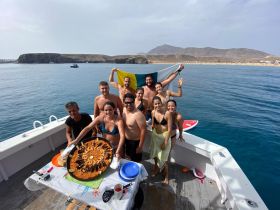 A lively group enjoys a delicious paella on board, celebrating together against a stunning coastal backdrop — perfect for memorable boat parties.