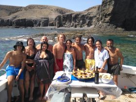 A joyful group poses for a photo on a boat, enjoying a delicious seafood feast against a stunning coastal backdrop — perfect for family gatherings and beach celebrations.