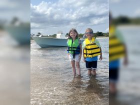Two children in life jackets stand in shallow water with a boat in the background, capturing a fun day at the beach — perfect for family adventures on the water.