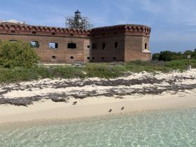 A view of a historic structure near the beach, with some vegetation and sandy shoreline visible. The image lacks clarity and detail.