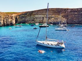 A picturesque scene of a sailboat anchored in crystal-clear waters, with people enjoying a swim nearby — a perfect representation of summer fun and relaxation on the water.