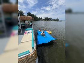 A fun group enjoys a sunny day on the water, lounging on a floating mat beside a pontoon boat — perfect for summer parties and relaxation.