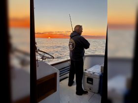 A fisherman stands on the deck of a boat, enjoying a stunning sunset over the ocean — a perfect moment for fishing enthusiasts.