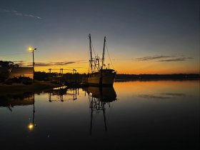 A serene sunset view of a fishing boat docked, reflecting beautifully on calm waters — a perfect scene for maritime enthusiasts and photographers.