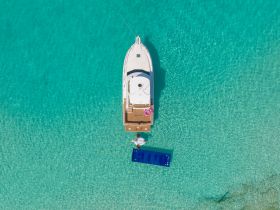 An aerial view of a boat anchored in crystal-clear waters, with a person enjoying a floating mat — a perfect scene for summer fun and relaxation on the water.
