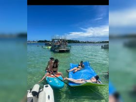 A lively group enjoys a fun day on the water, paddleboarding and relaxing on floating mats, creating the perfect summer party atmosphere.